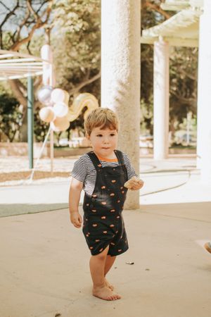 Small child wearing black overalls with hearts walking on a concrete path under white columns.