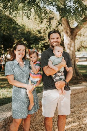 Family photoshoot in a park with trees casting dappled sunlight on a warm summer day.