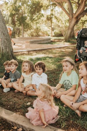 Children sitting together outdoors on a sunny day under trees.