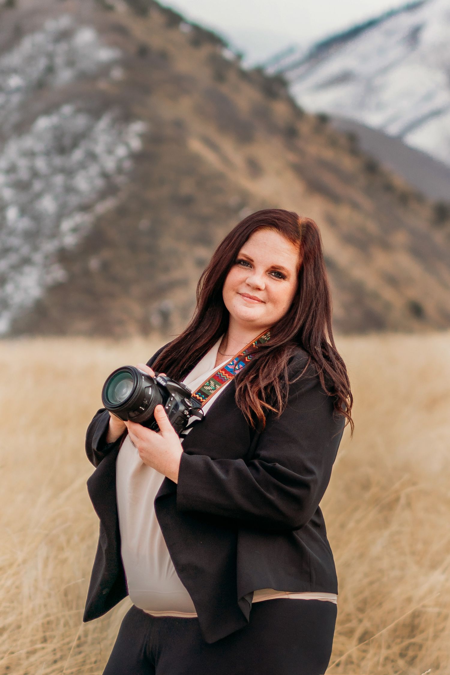A photographer holding a professional camera poses in a natural outdoor setting with mountains in background.