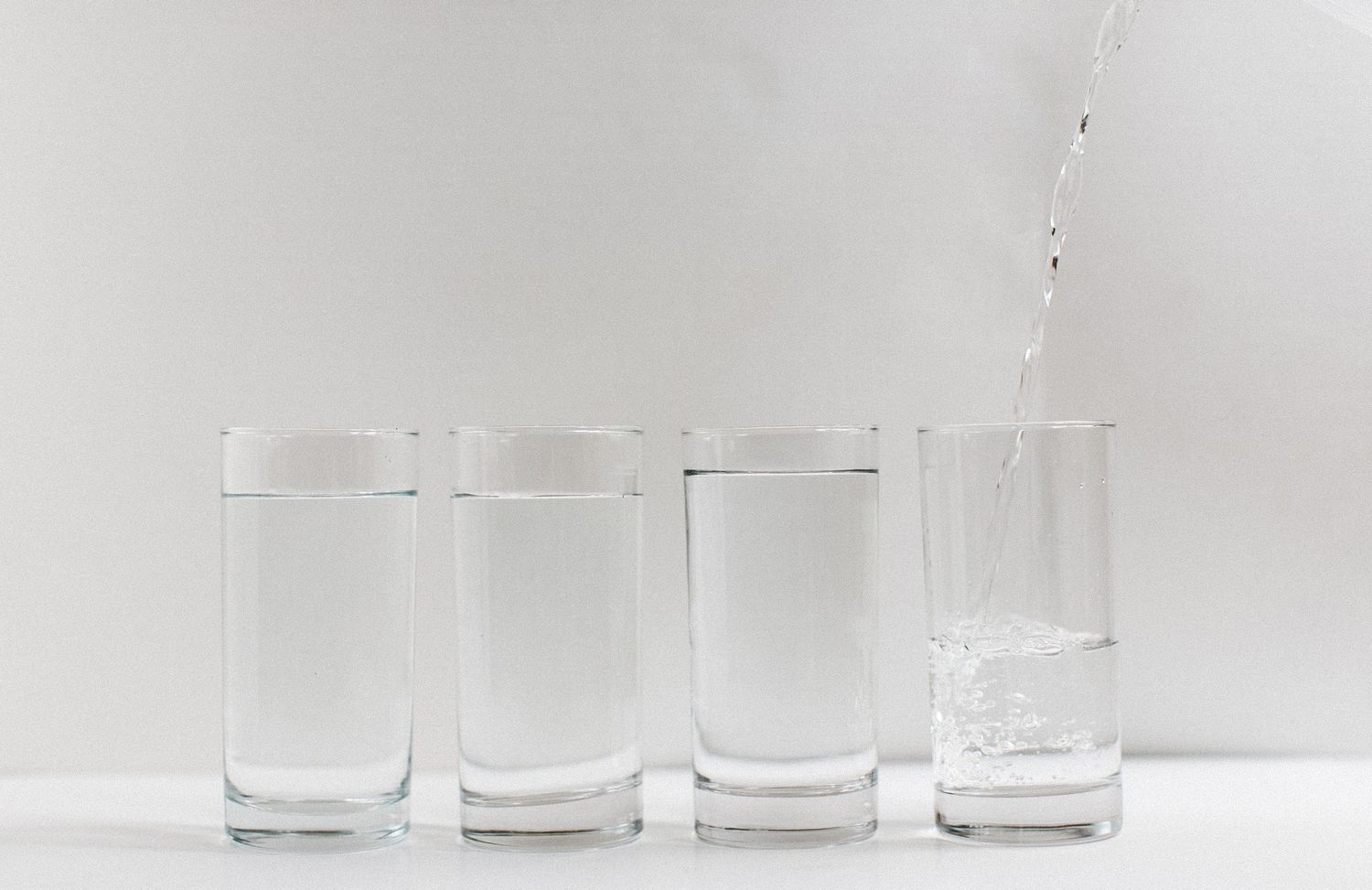 Line of clear drinking glasses with water being poured into the last glass on white background.