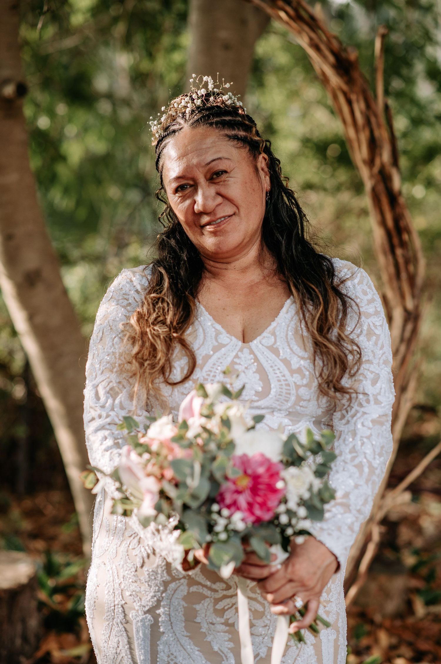 Bride in lace gown and floral crown holds colorful bouquet against natural garden backdrop.