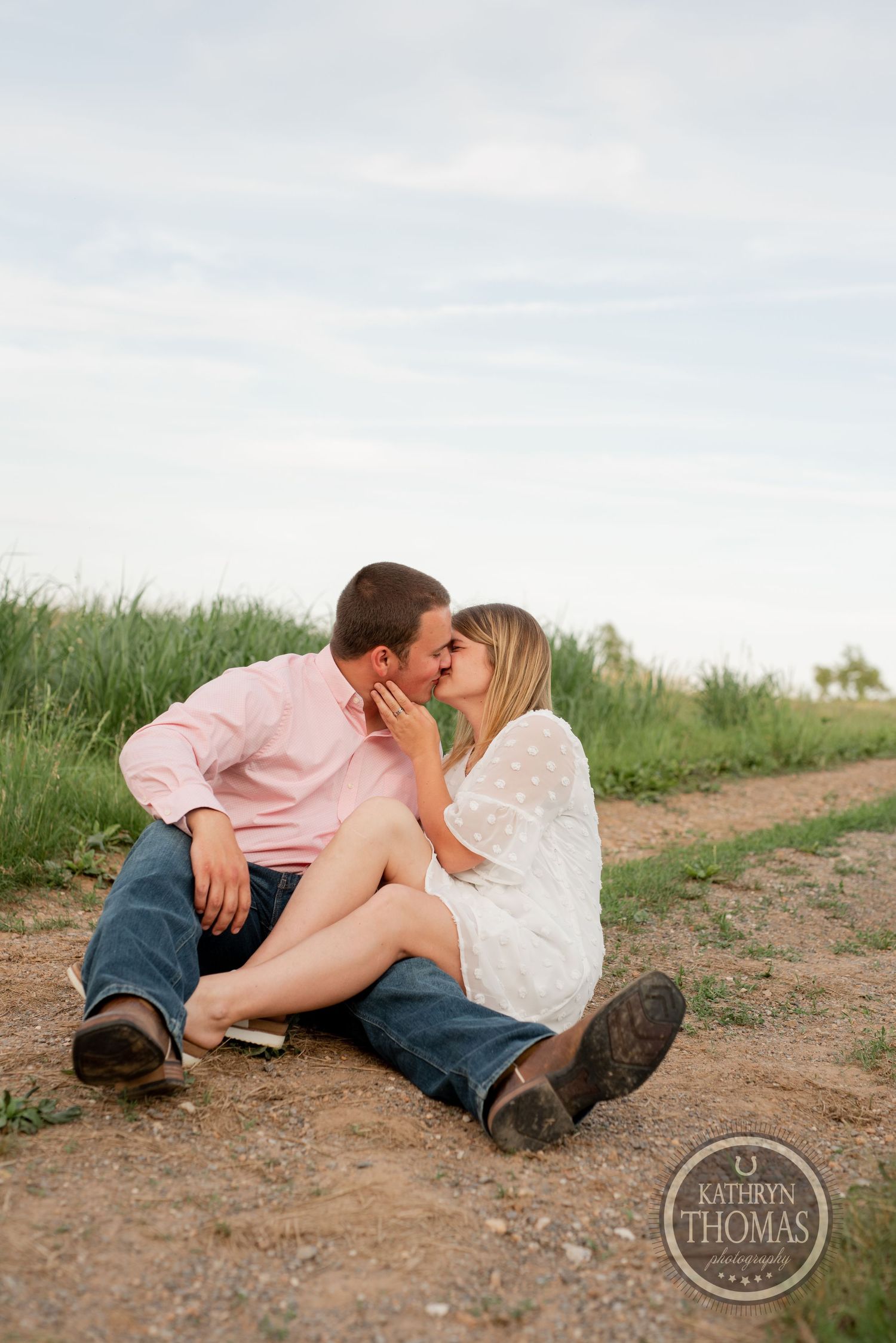 A couple shares a tender moment while sitting on a dirt path surrounded by grass.