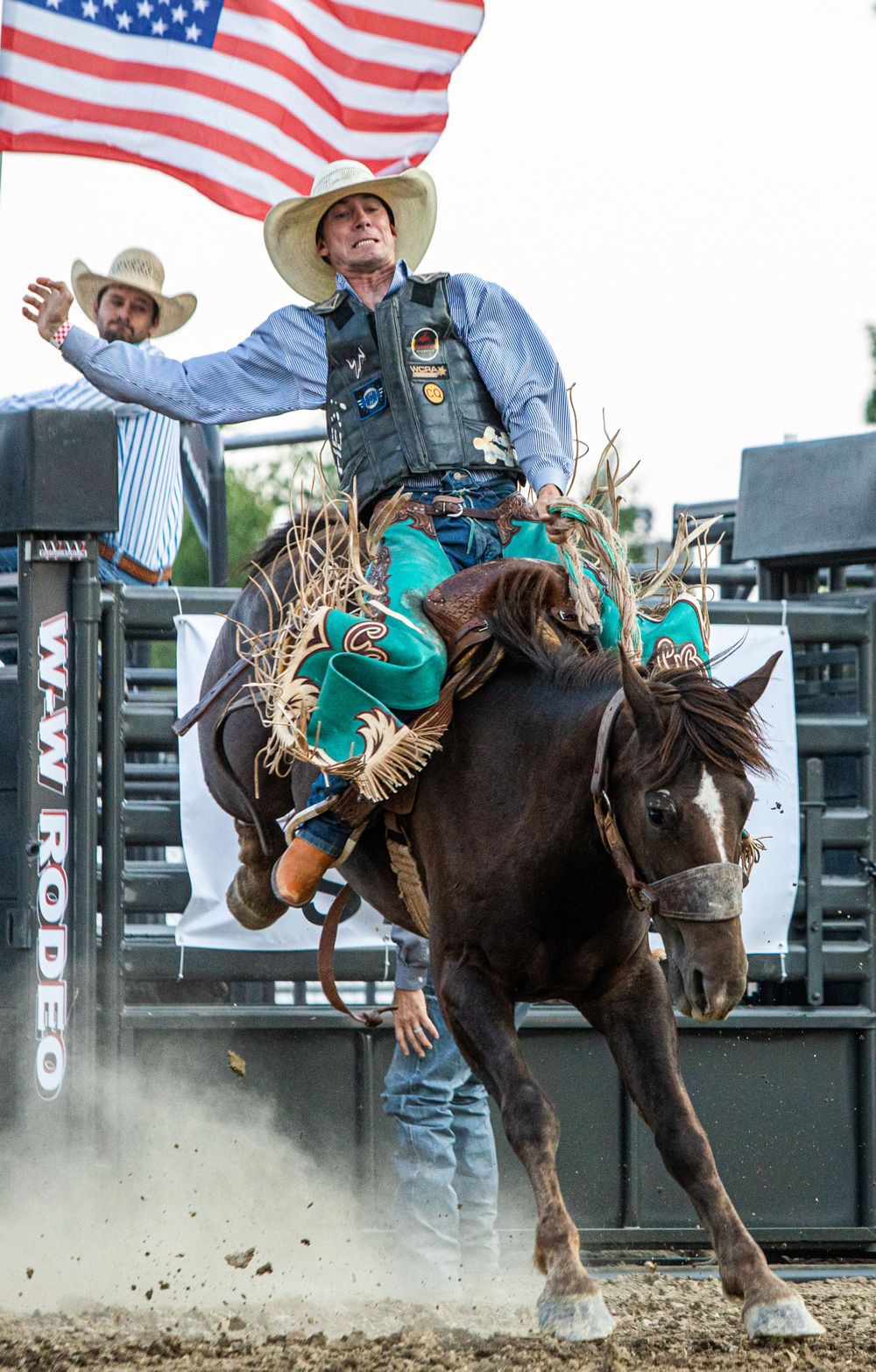 Rodeo rider on bucking horse with American flag waving in background.