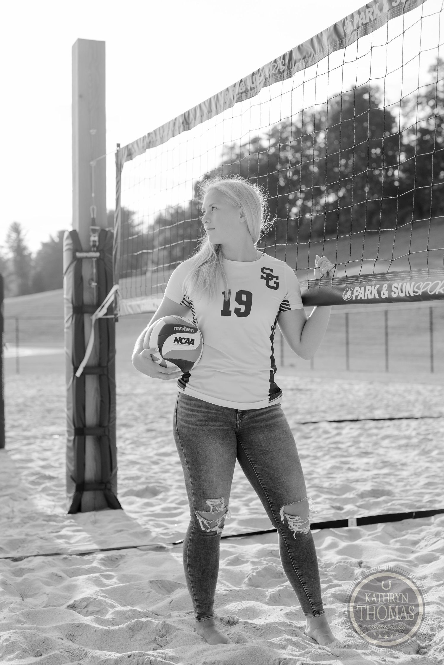 A black and white photo of an athlete posing with a volleyball by the court net.