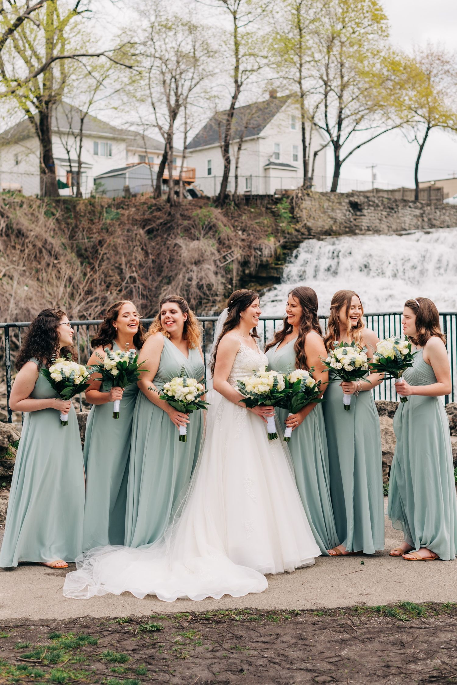 bride and bridesmaids in eucalyptus green dresses pose in front of a waterfall