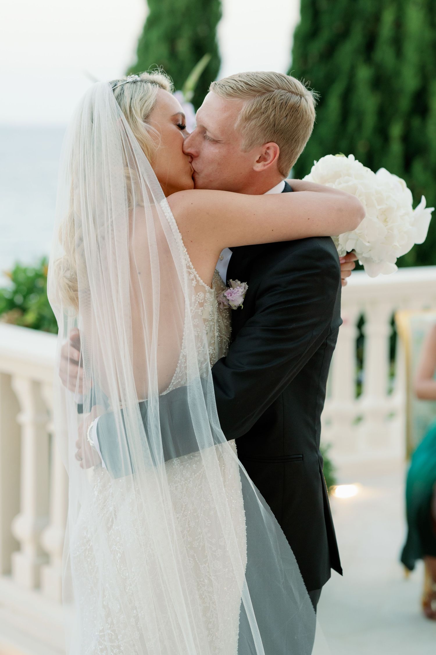 Romantic kiss between newlyweds with flowing veil against cypress tree backdrop.