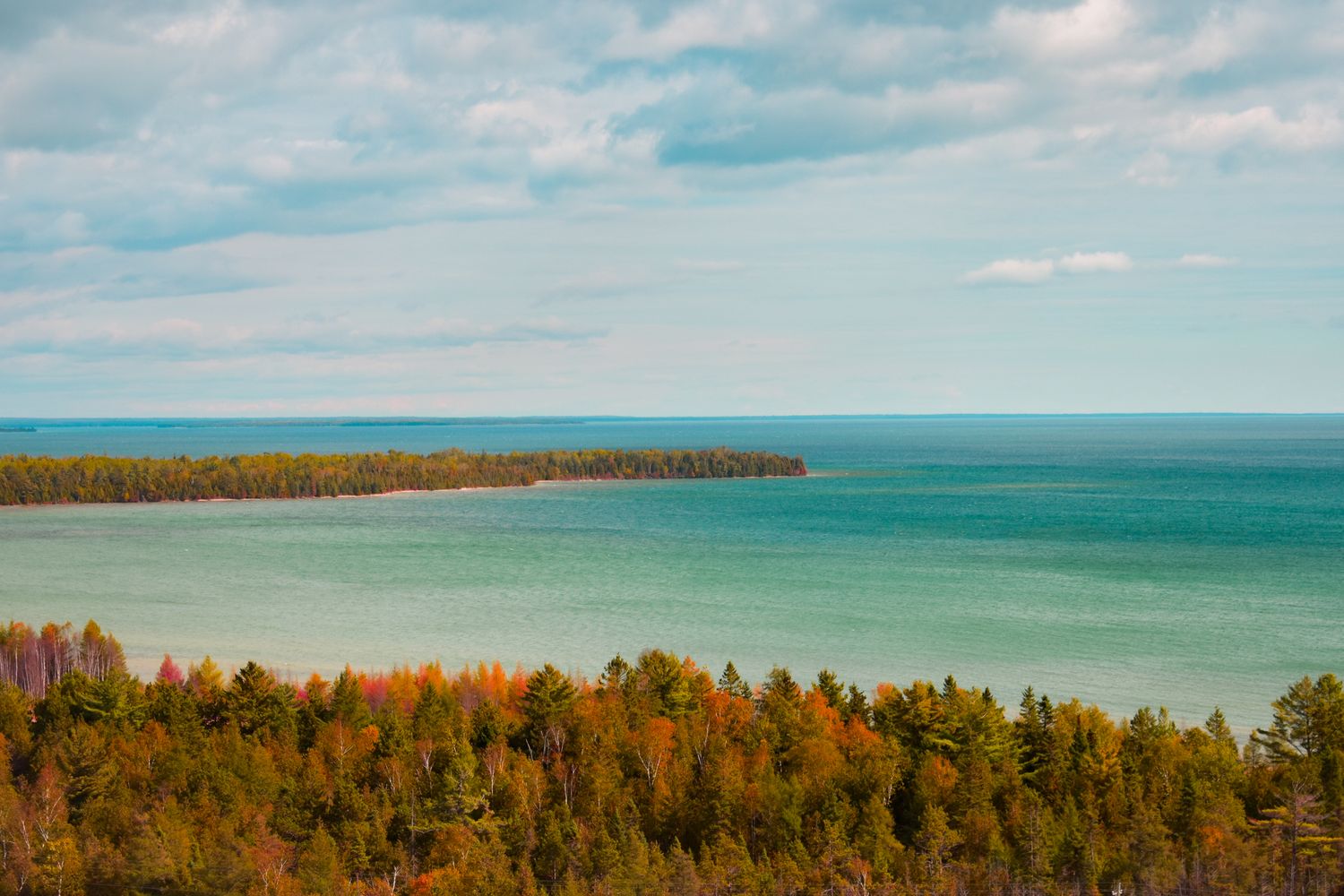 Aerial view of a turquoise coastline with autumn trees and a curved peninsula stretching into the blue ocean.