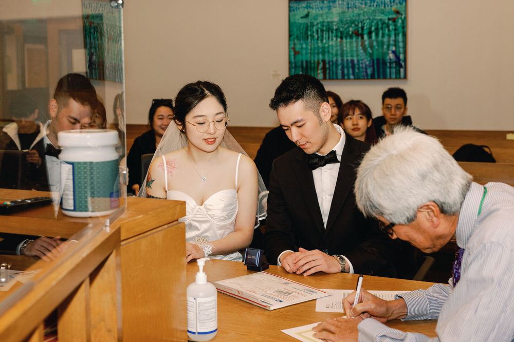 Asian bride and groom signing marriage documents at a courthouse counter with guests watching.
