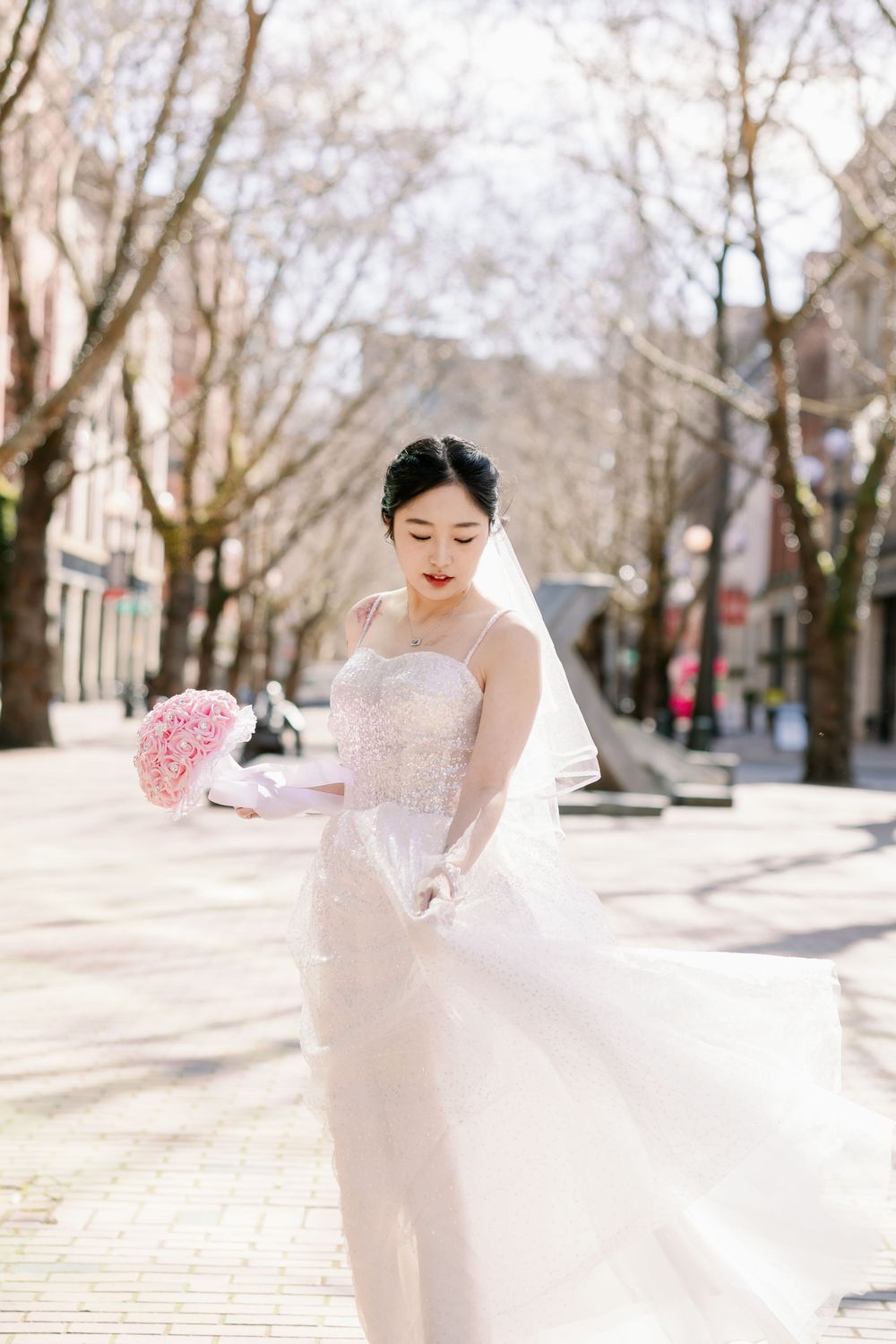 Elegant bride in beaded white gown and veil holds pink bouquet on sunny tree-lined street.