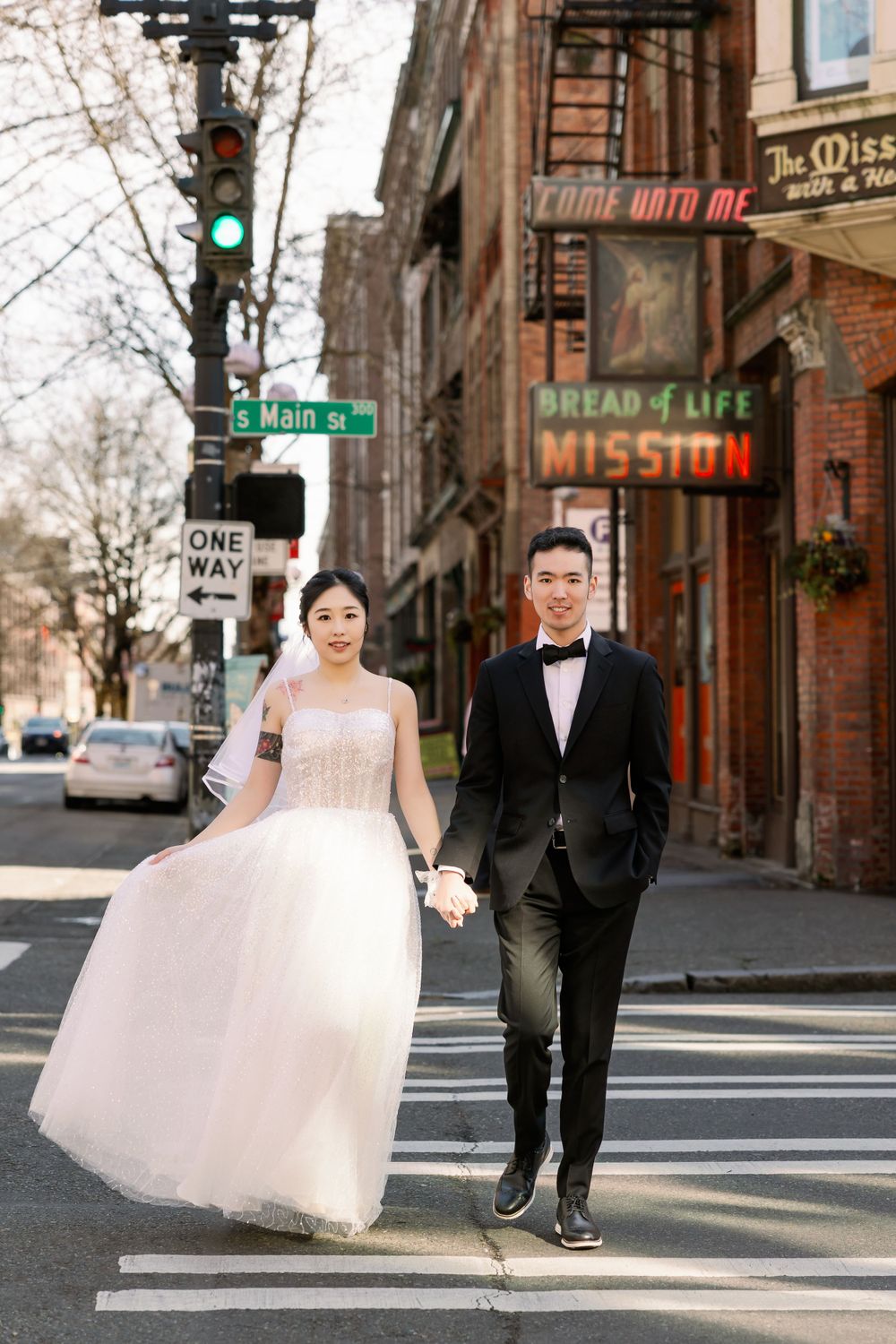 Asian wedding couple crossing a city street near S Main St sign, bride in white gown and groom in black tuxedo.