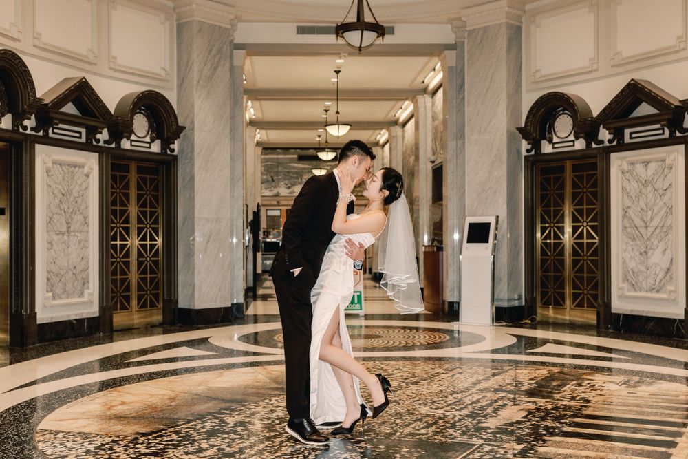 Asian groom lifting his bride for a romantic kiss in a luxurious hotel lobby with marble floors.
