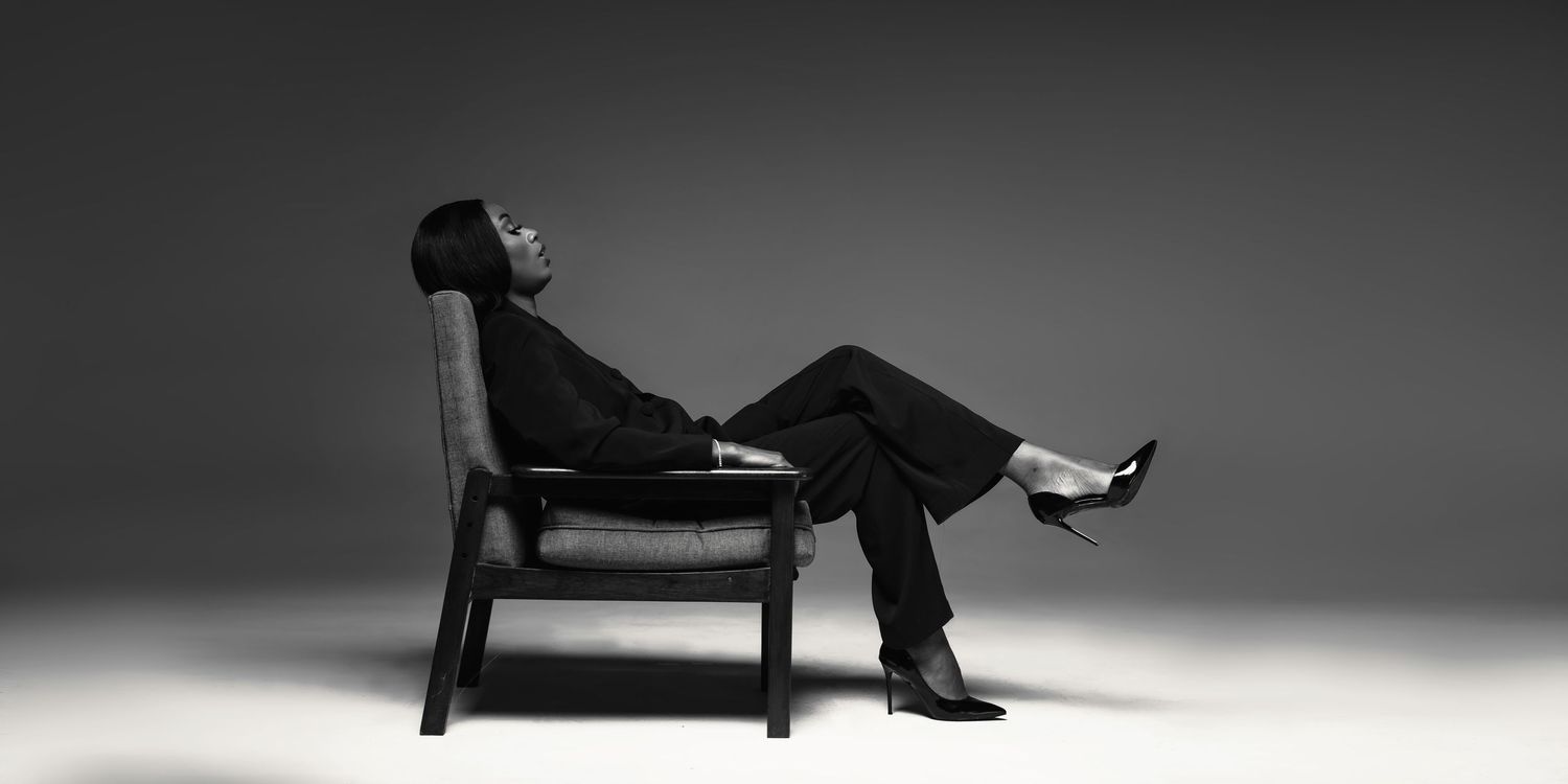 Artistic black and white portrait of someone reclining in a wooden chair against dark background.