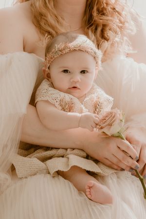 Intimate portrait of baby wearing delicate lace dress and headband holding rose against white tulle background.