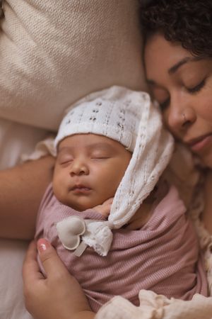 Sleeping baby wearing pink outfit and white knit hat rests peacefully in loving embrace.