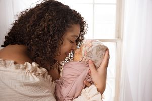Tender moment with a sleeping newborn baby in pink clothing against a bright window backdrop.
