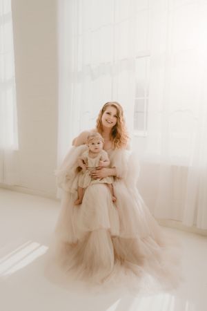 Person in flowing tulle gown holds baby wearing matching dress in bright white room.