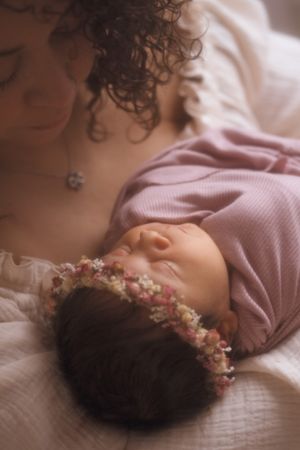 Newborn baby wearing floral crown and pink outfit sleeping peacefully on white bedding.