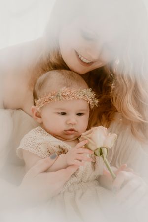 Ethereal portrait of baby in lace dress and gold headband holding a pink rose with soft dreamy vignette effect.