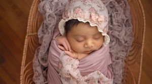 Sleeping newborn wearing lace bonnet and wrapped in pink fabric on vintage chair.
