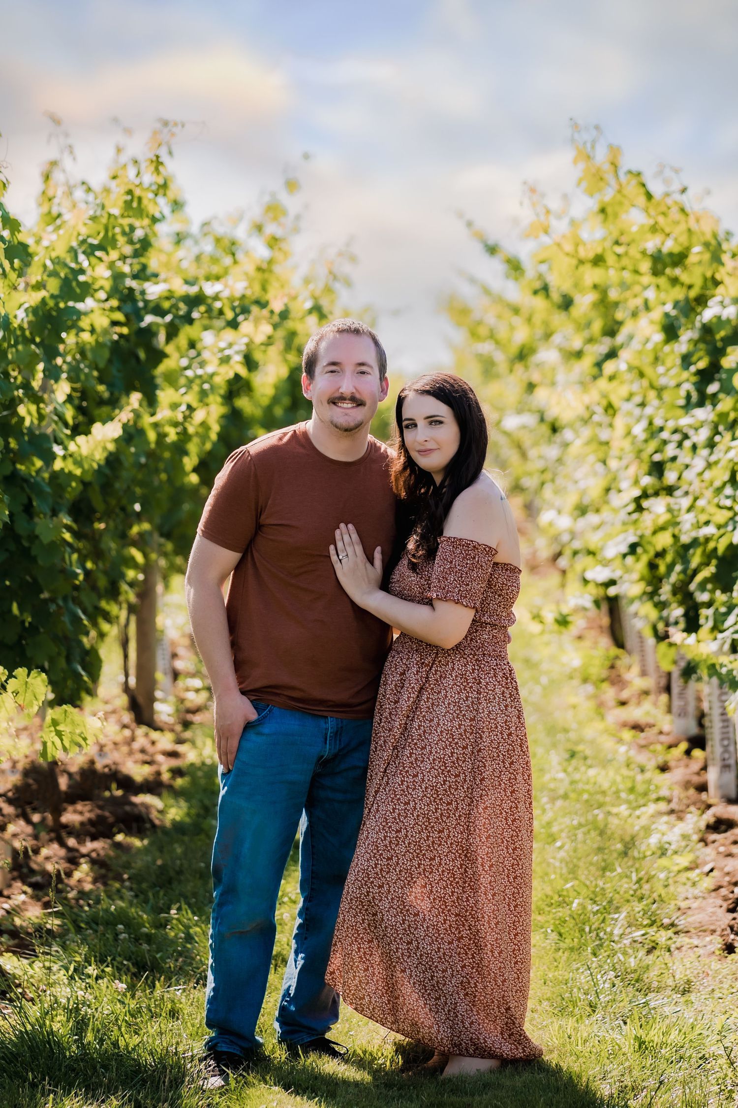 Couple standing together in sunny vineyard with rows of grape vines.