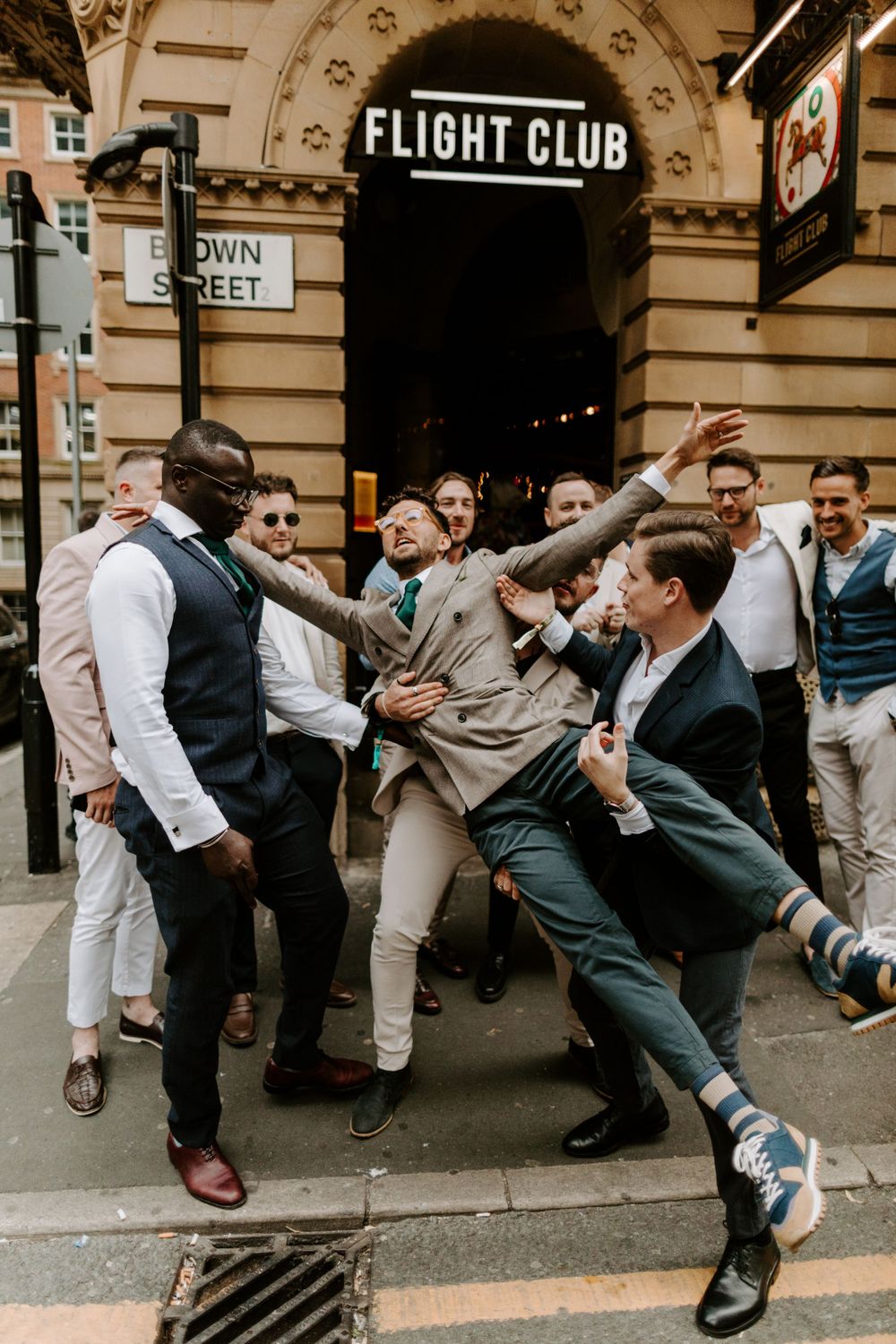 A lively group of groomsmen posing playfully outside Flight Club in an urban setting.