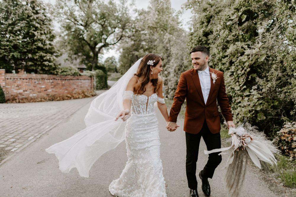 Couple walking hand in hand with flowing veil and rustic bouquet on stone path.