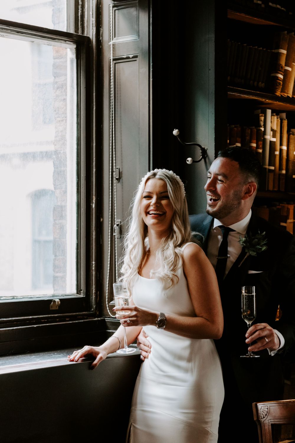 Couple shares laughs by window during wedding celebration with champagne.