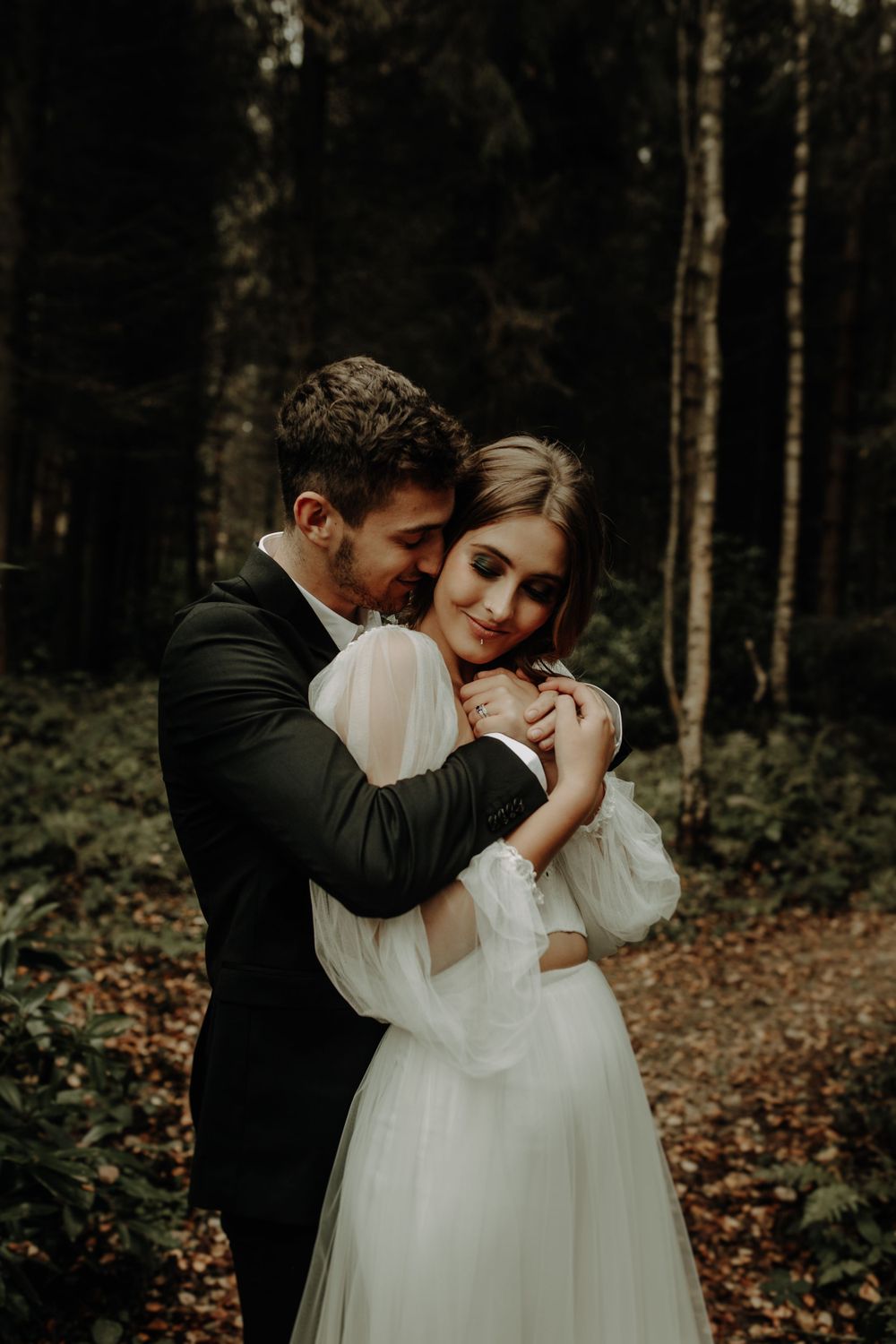 A romantic embrace between a couple in wedding attire among tall forest trees.