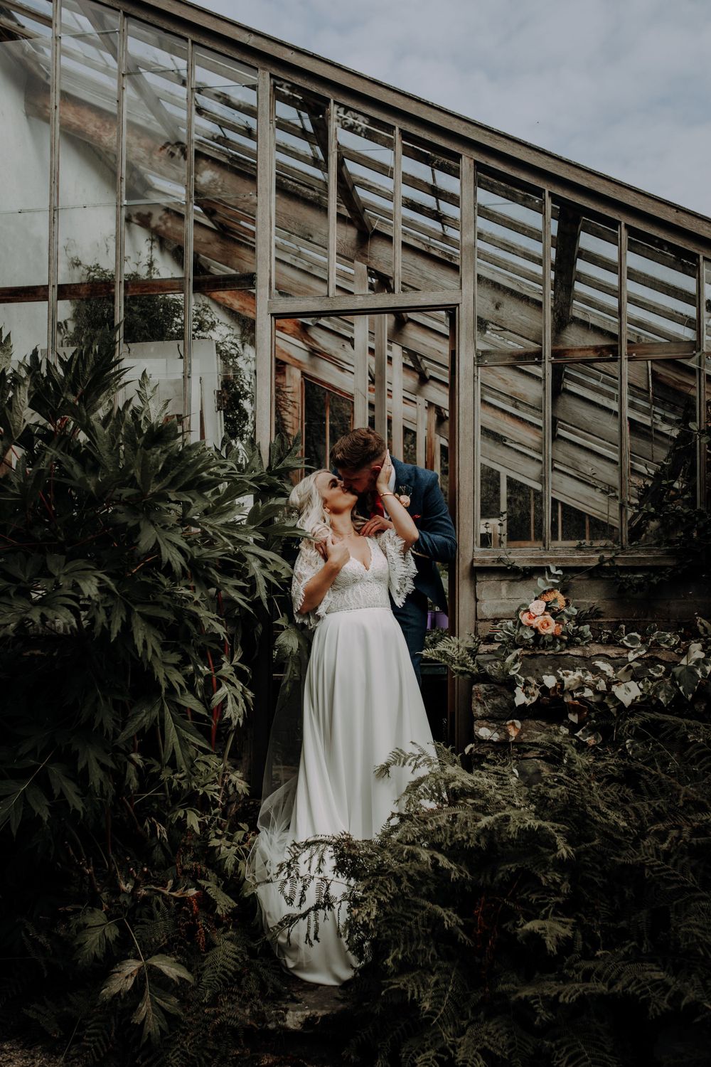 A couple shares a tender moment in front of a greenhouse filled with lush plants.