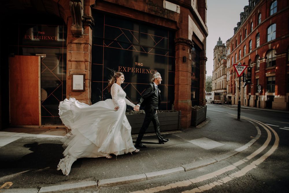 A bride's dress flows dramatically on a curved street corner in an urban setting.