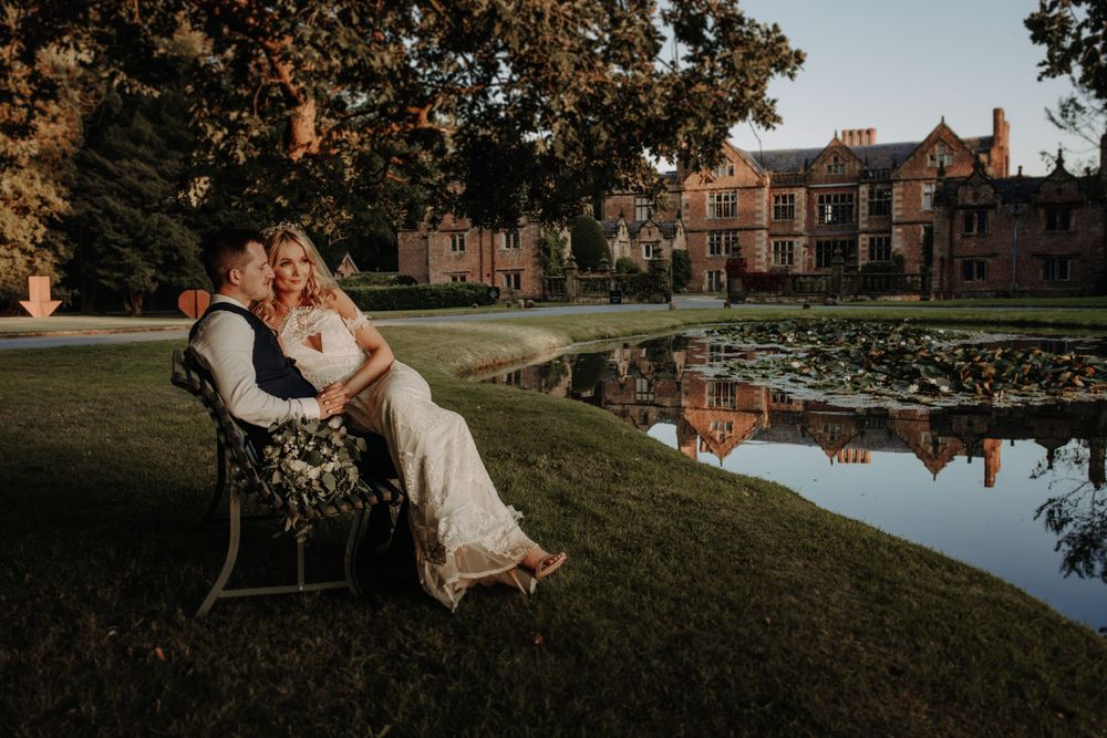 A couple shares an intimate moment on a bench near a reflective pond with a grand estate in the background.