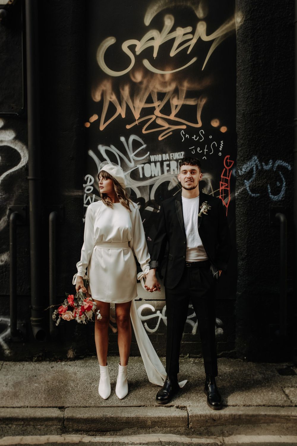 A fashionable couple poses against an urban graffiti wall in wedding attire.