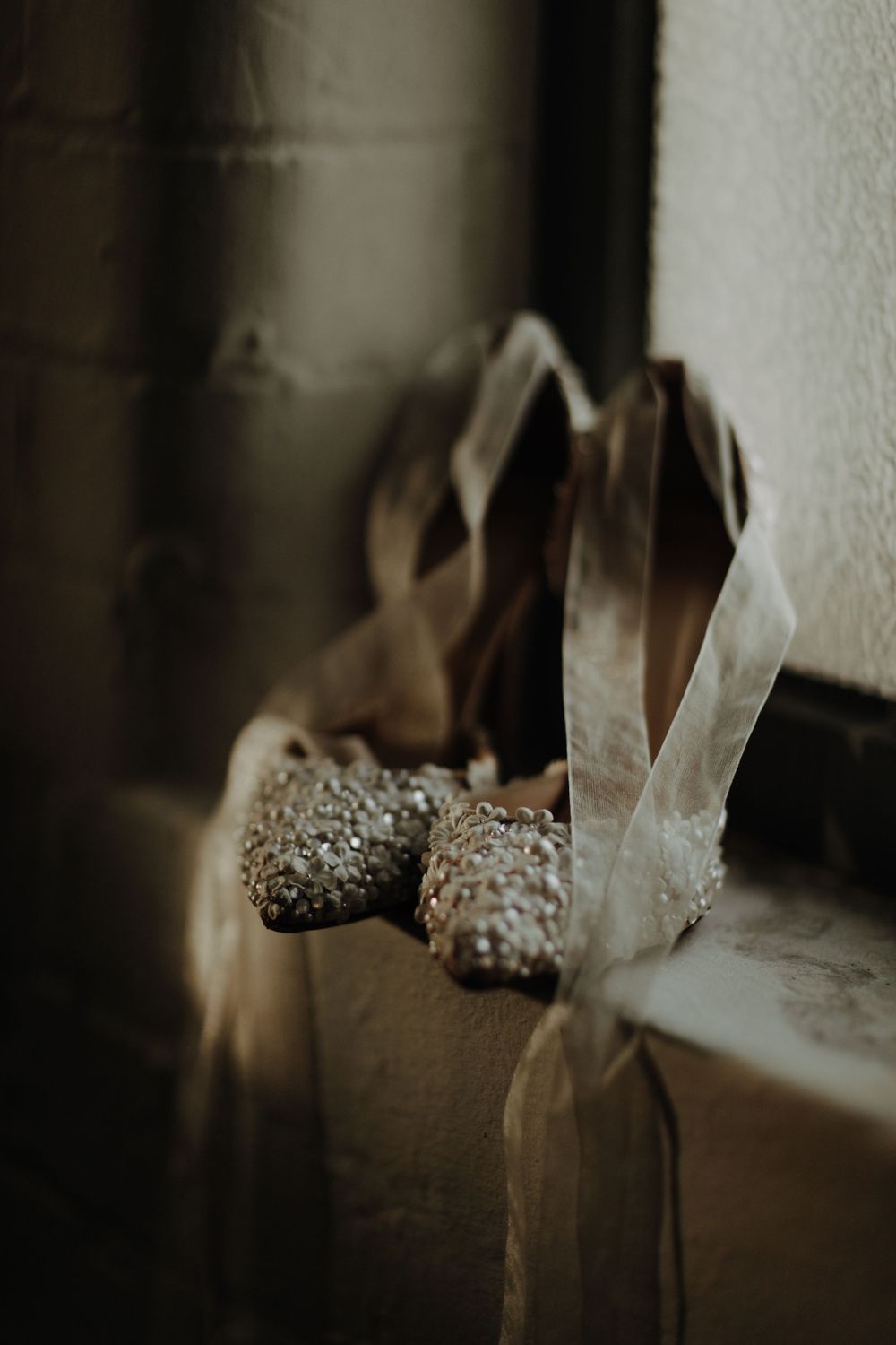 Vintage sparkly bridal shoes sit on a windowsill in moody lighting.