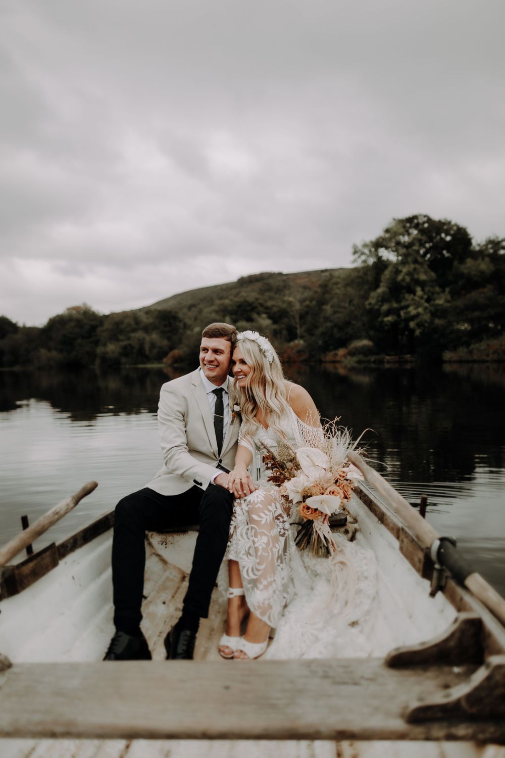 A couple sits together in a wooden boat on a serene lake surrounded by hills.