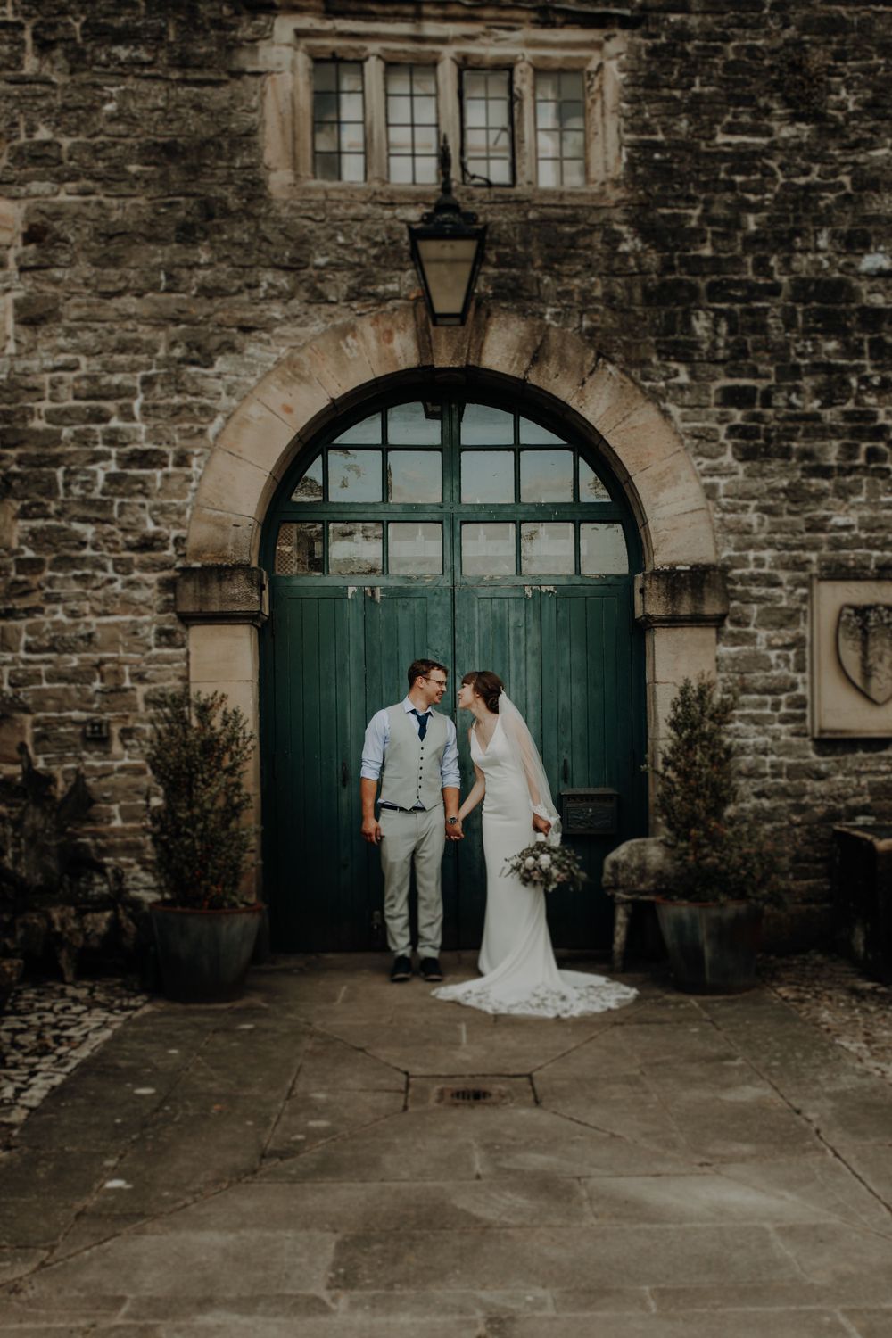 A romantic wedding couple poses in front of a rustic green door and stone wall with an arched entrance.