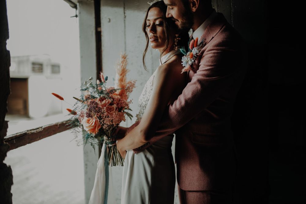 A couple in wedding attire share an intimate moment by a window with a coral-colored bouquet.
