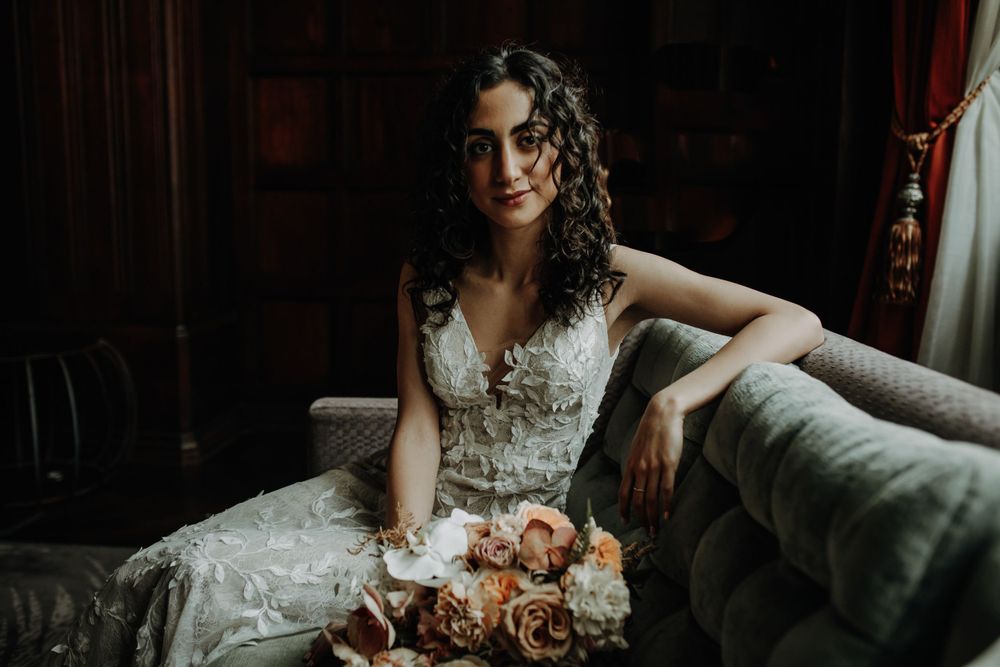 Bride in detailed lace dress sits elegantly on vintage couch in moody lighting.