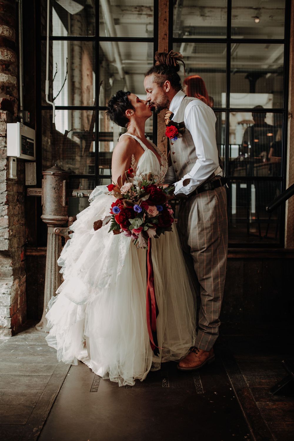 Couple in vintage-inspired wedding attire embrace near industrial windows holding vibrant bouquet.