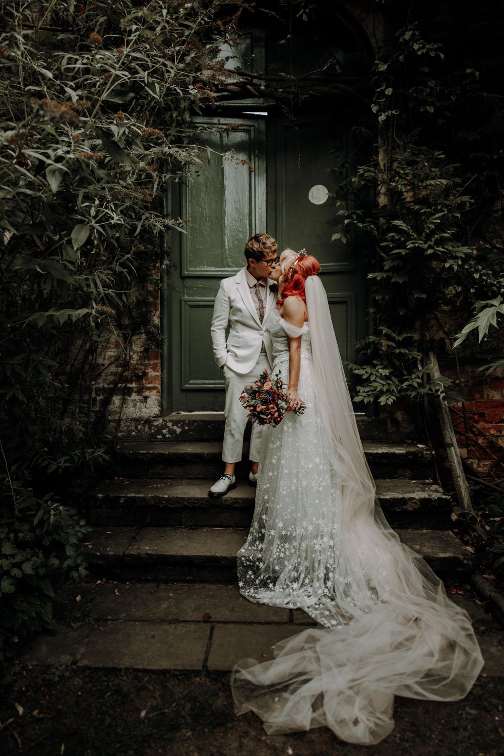 A couple stands on stone steps against a dark green door in vintage-style wedding attire.
