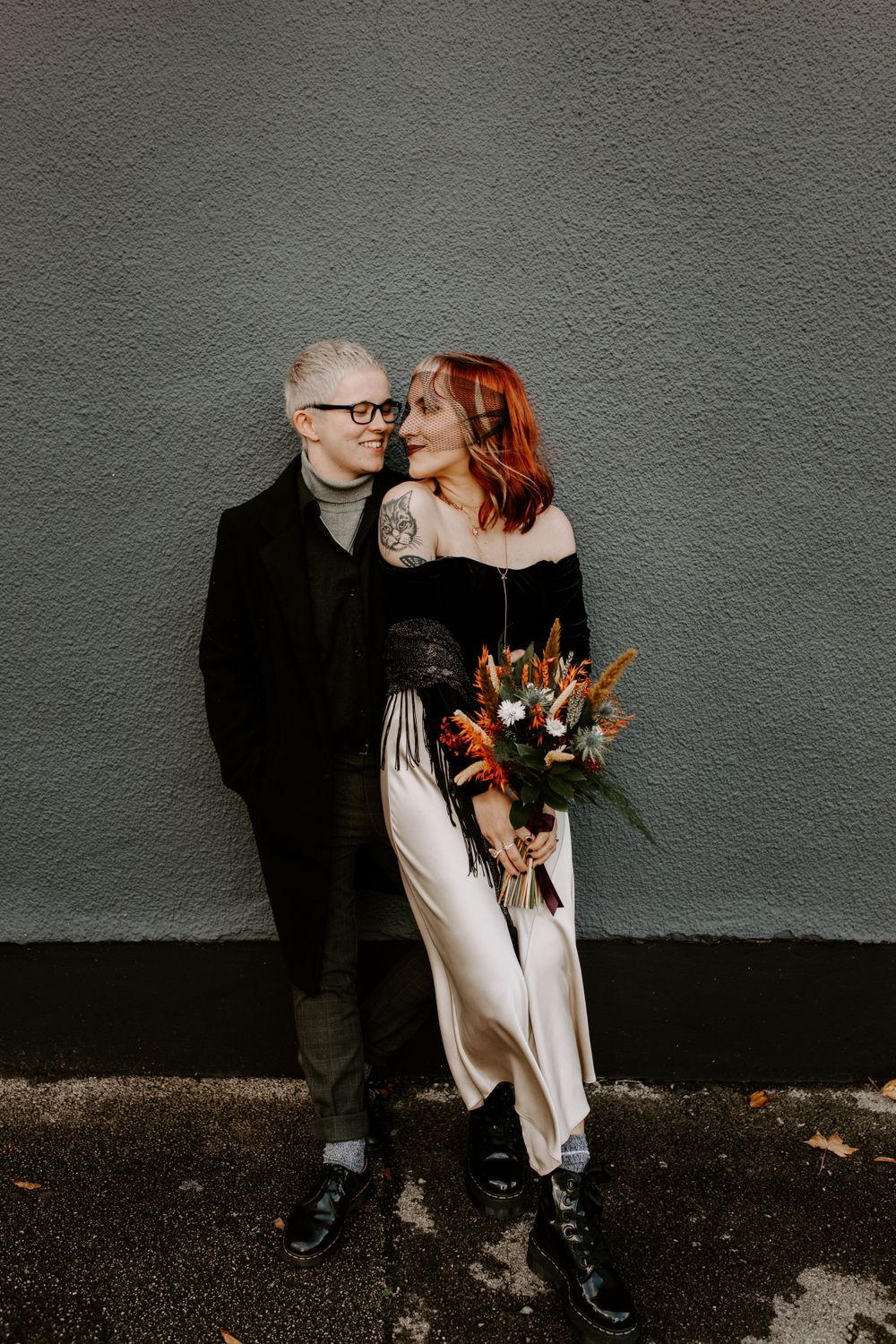 A couple in contrasting black and white outfits pose against a grey wall with a rustic bouquet.