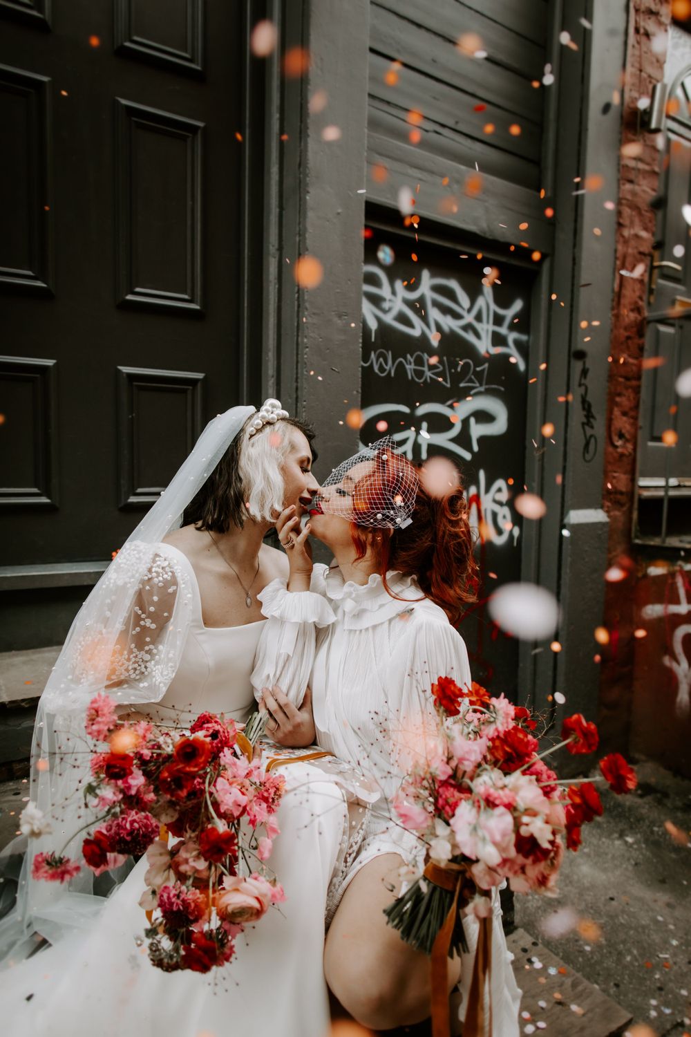 Two brides in white lace dresses celebrating with confetti and red floral bouquets against black doors.