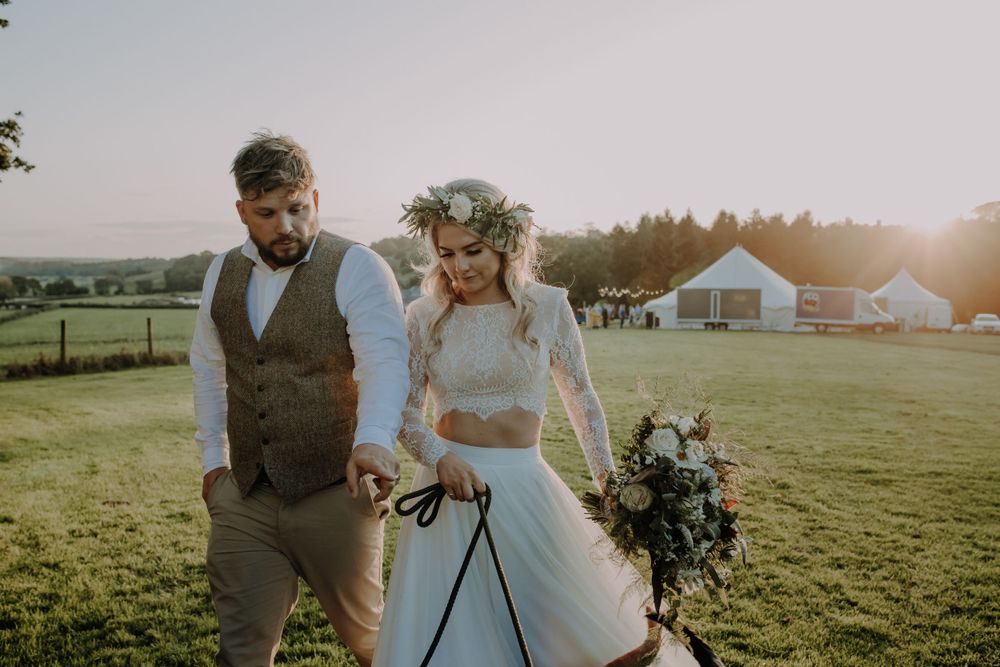 A couple walks through a sunlit meadow with a flower crown and rustic wedding attire.
