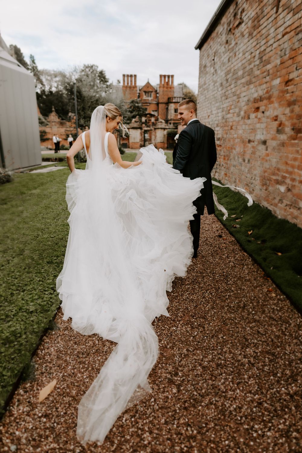 Long flowing wedding dress train sweeps across pathway near historic brick building.