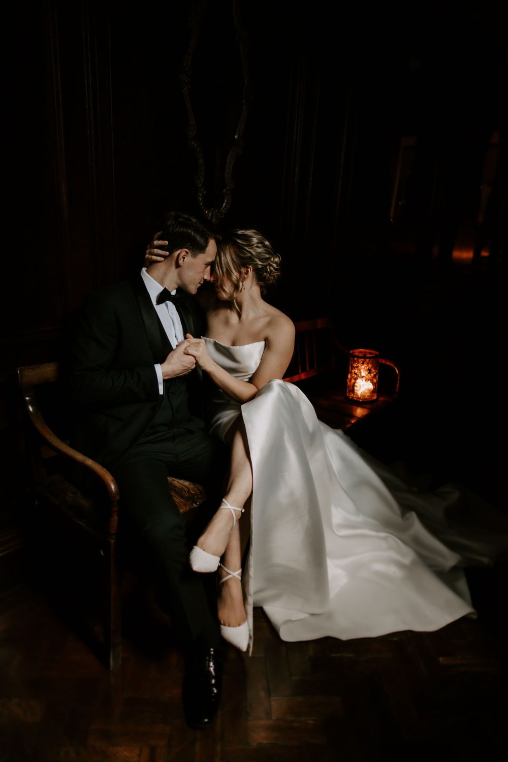 Romantic wedding couple sharing an intimate moment in formal attire by candlelight in a dark moody setting.