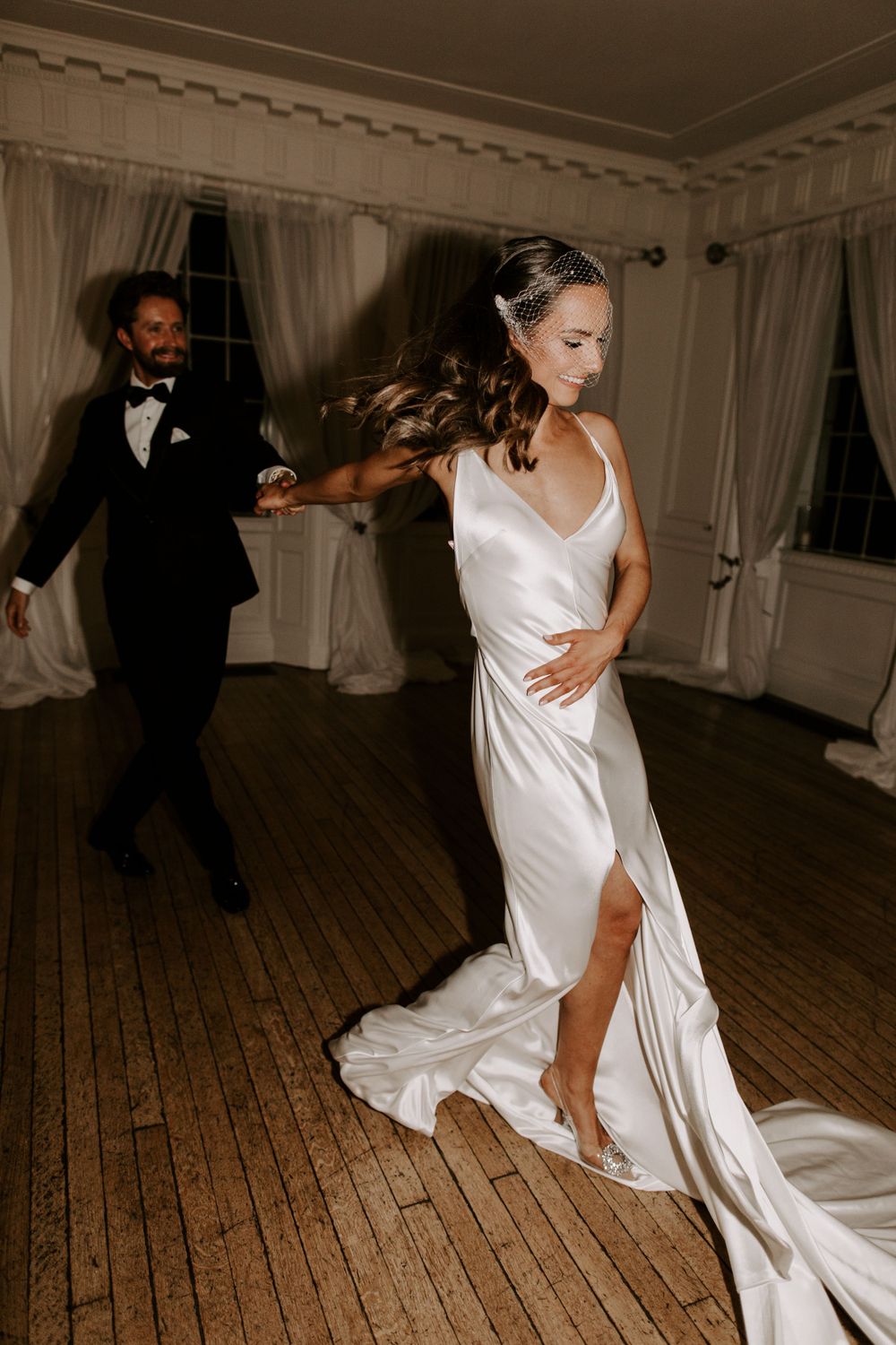 A couple dances in an elegant room with wooden floors and white curtains.