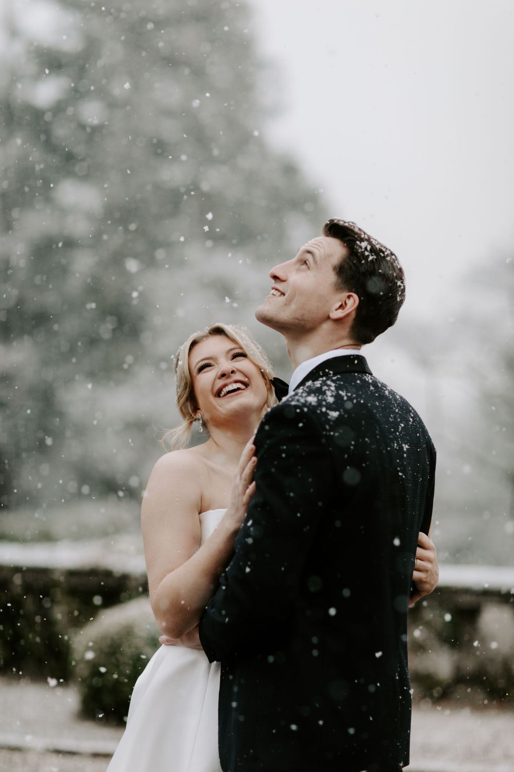 A couple laughs joyfully together in the falling snow during their winter wedding photoshoot.