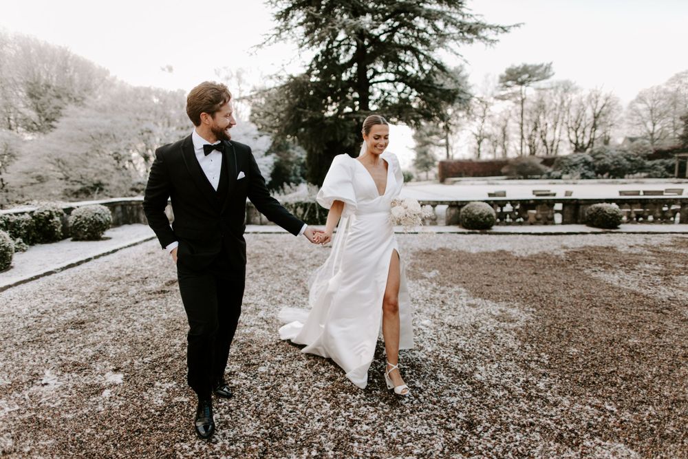 A couple walks hand in hand through a garden estate in wedding attire.