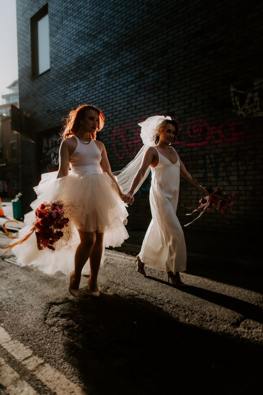 Two friends dance in white dresses on an urban street at sunset, one carrying a bouquet.