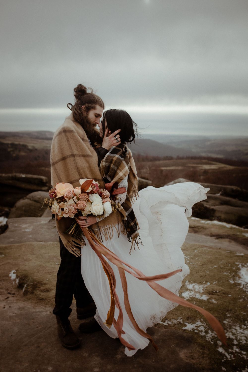 A romantic wedding couple embraces on a mountaintop overlooking valleys while holding a floral bouquet.